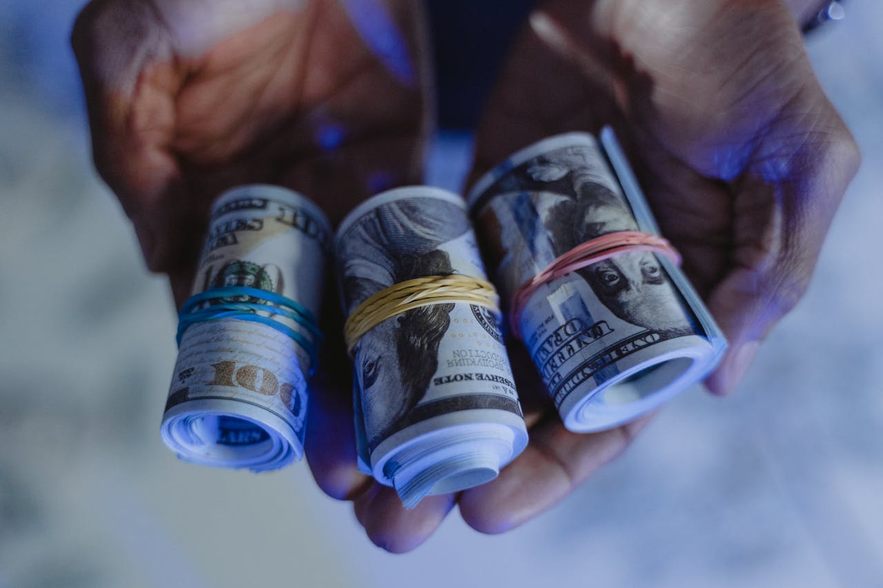 Close-up of hands holding rolled US dollar banknotes secured with rubber bands, set against a blue-lit background.